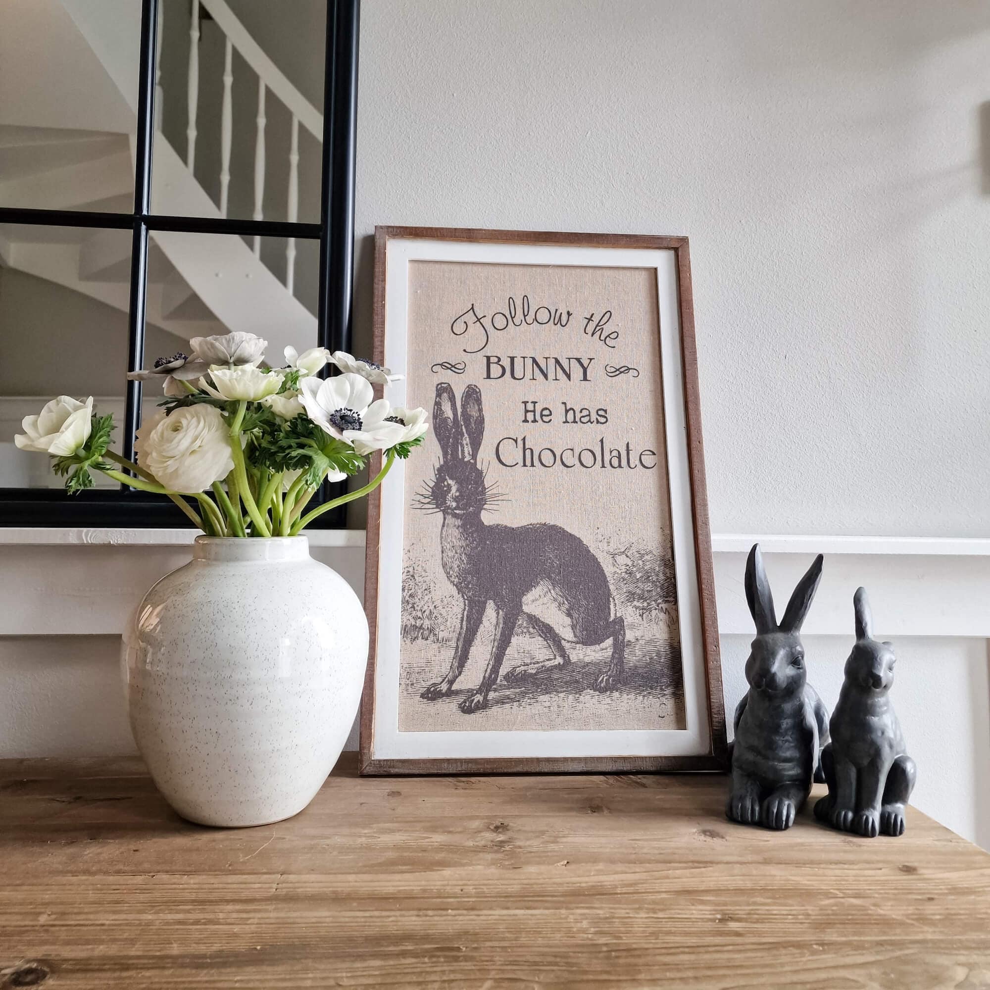 Quadro decorativo con coniglio e testo, fiori bianchi in vaso di ceramica, due statuette di conigli su tavolo in legno