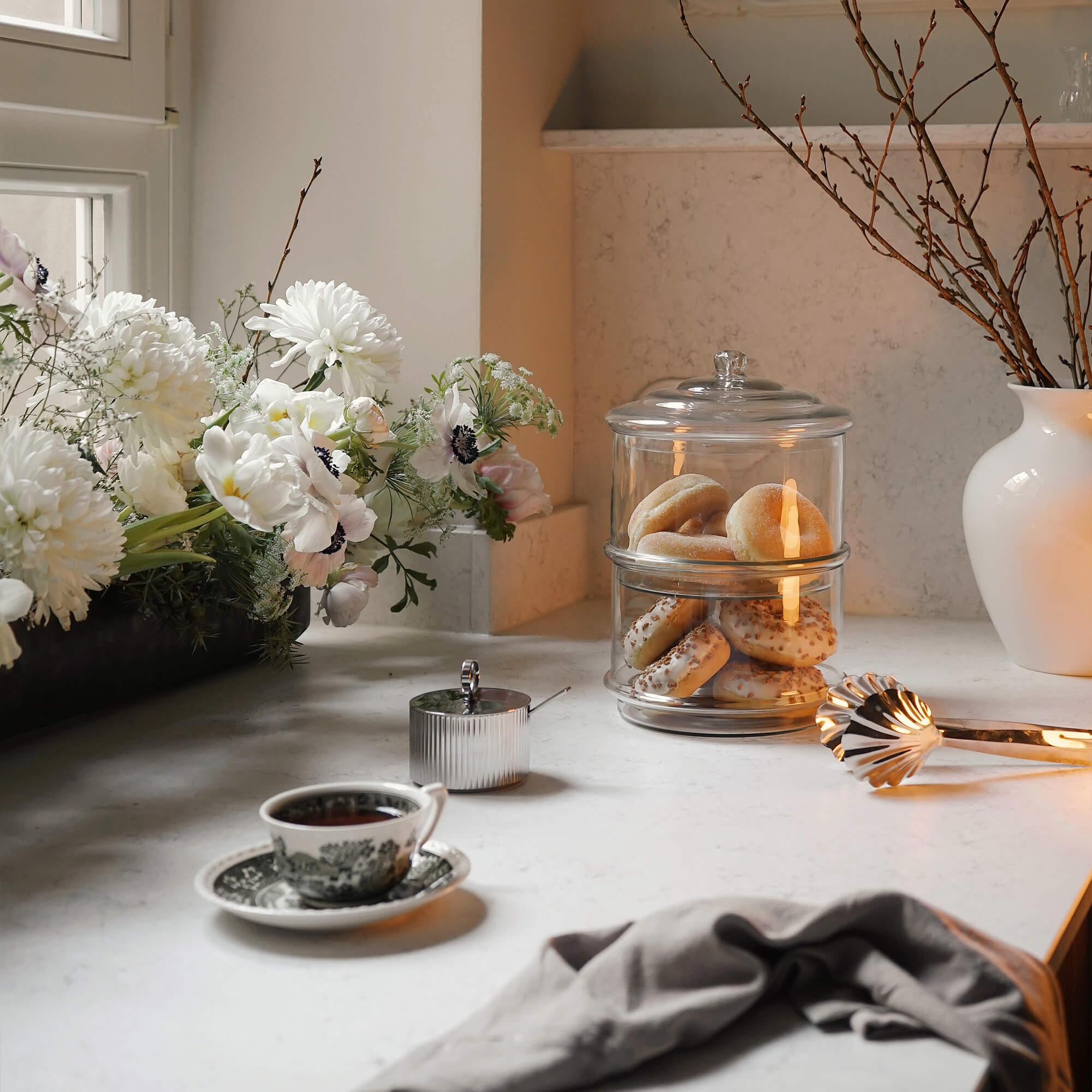 Contenitore in vetro a due piani per biscotti su piano cucina chiaro con fiori e tazza di tè