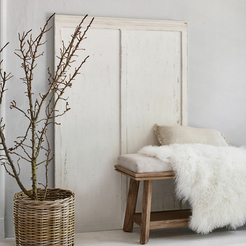 Panca rustica in legno con cuscino beige e coperta bianca in pelliccia sintetica accanto a un cesto di vimini con rami