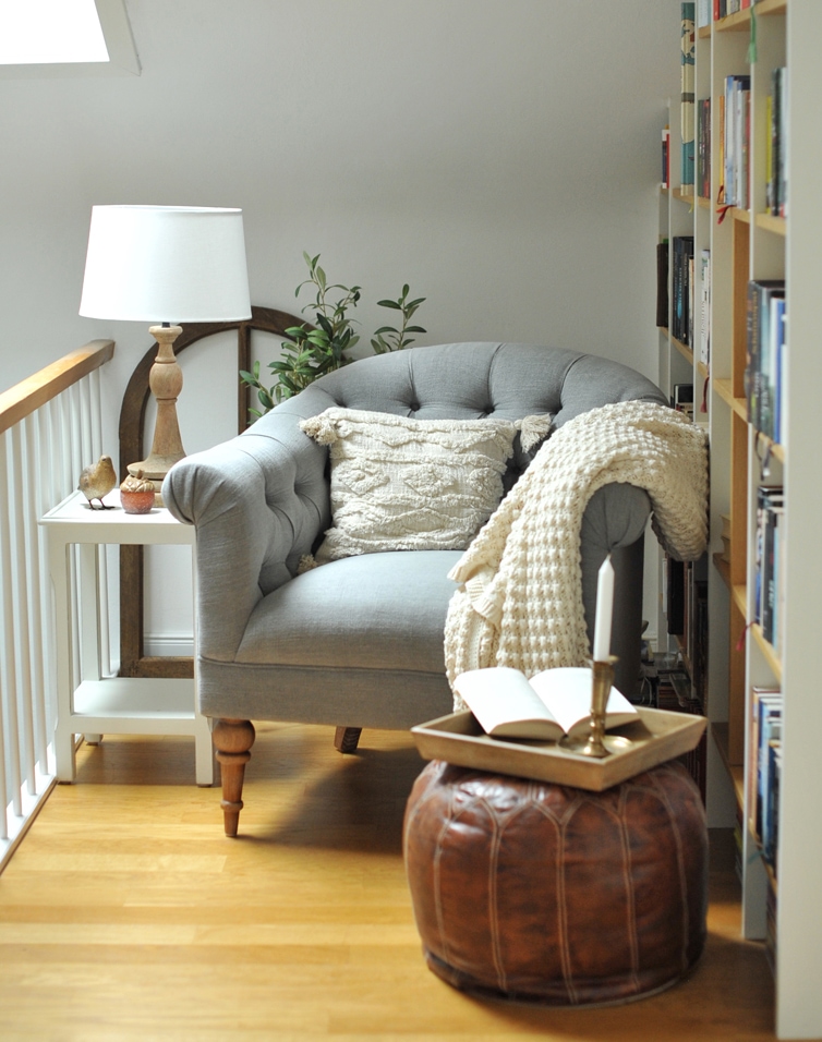 Divano grigio imbottito con gambe in legno, cuscino crema e coperta, pouf in pelle marrone e lampada su tavolo