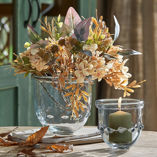 Vaso di vetro con fiori secchi beige e marroni e portacandela con candela su tavolo in legno