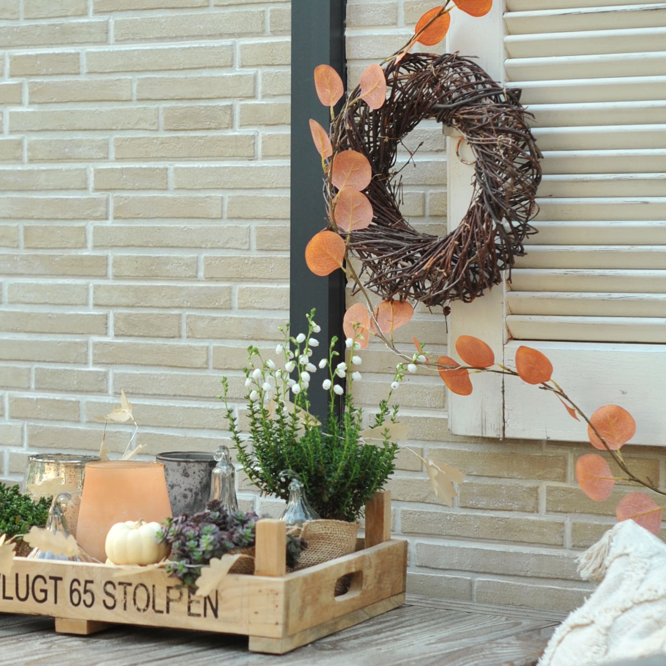Vassoio in legno con decorazione autunnale di piante verdi e fiori bianchi davanti a muro di mattoni