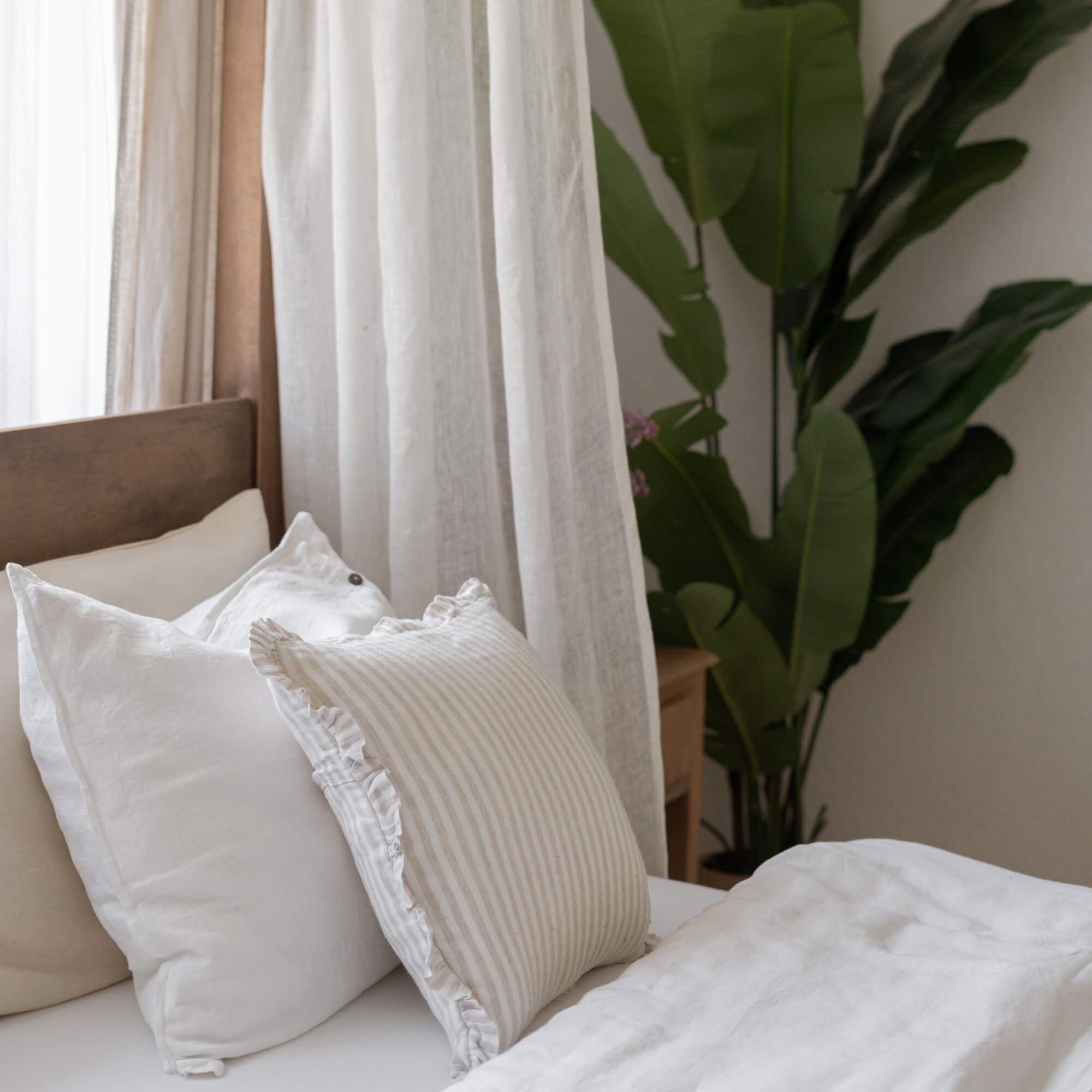 Cuscini in lino bianco e beige su letto con testiera in legno e pianta verde sullo sfondo