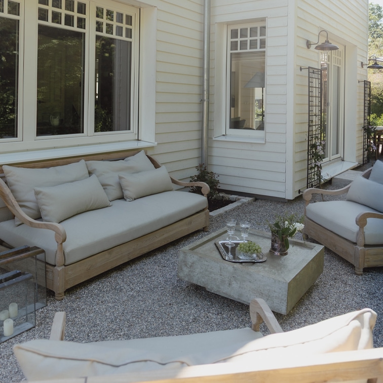Set di mobili da giardino in legno chiaro con cuscini grigi su patio ghiaioso davanti a casa bianca