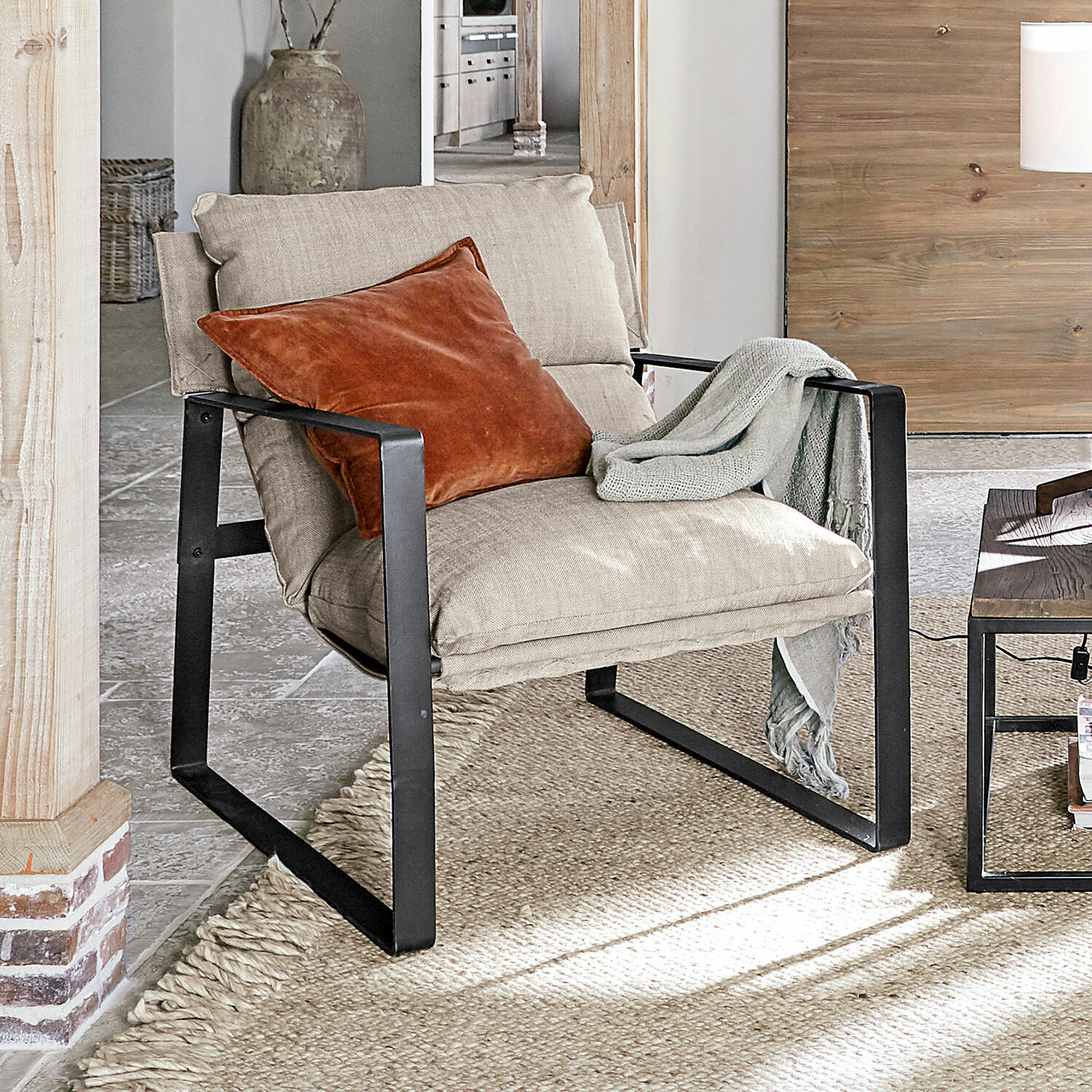 Poltrona Seuvard Poltrona in tessuto beige con struttura metallica nera, cuscino arancione e coperta grigia in soggiorno