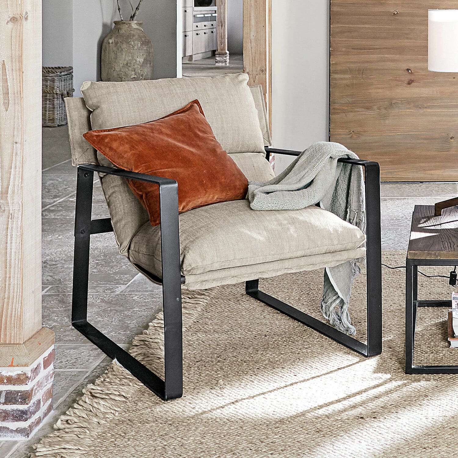 Poltrona Seuvard Poltrona in tessuto beige con struttura metallica nera, cuscino arancione e coperta grigia in soggiorno
