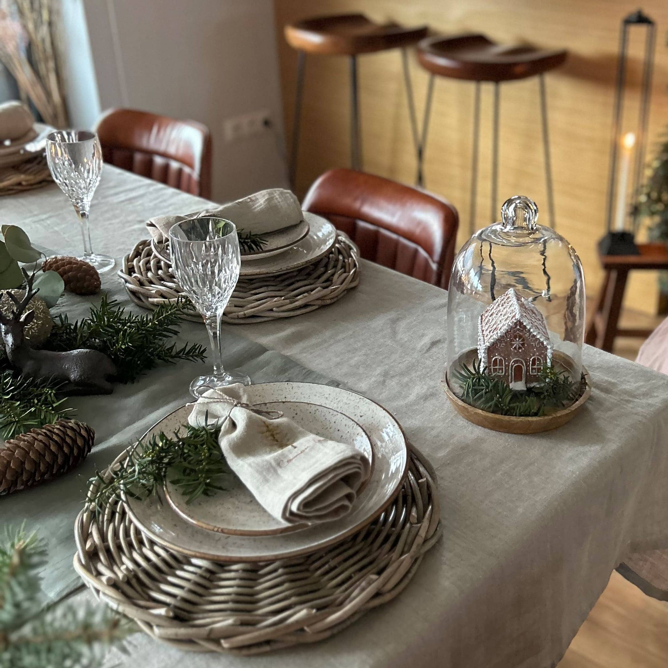 Tavola elegantemente apparecchiata con sottopiatti in vimini, piatti in ceramica e decorazione natalizia con casetta sotto campana di vetro