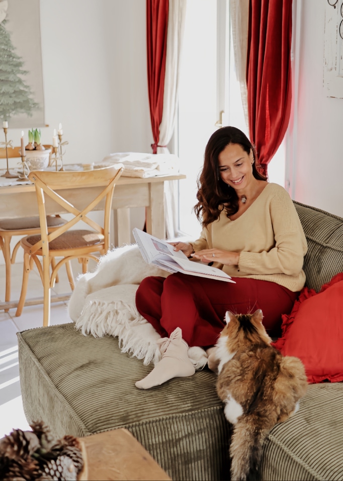 Donna con maglione beige e pantaloni rossi legge un libro su un divano verde con un gatto in un soggiorno accogliente