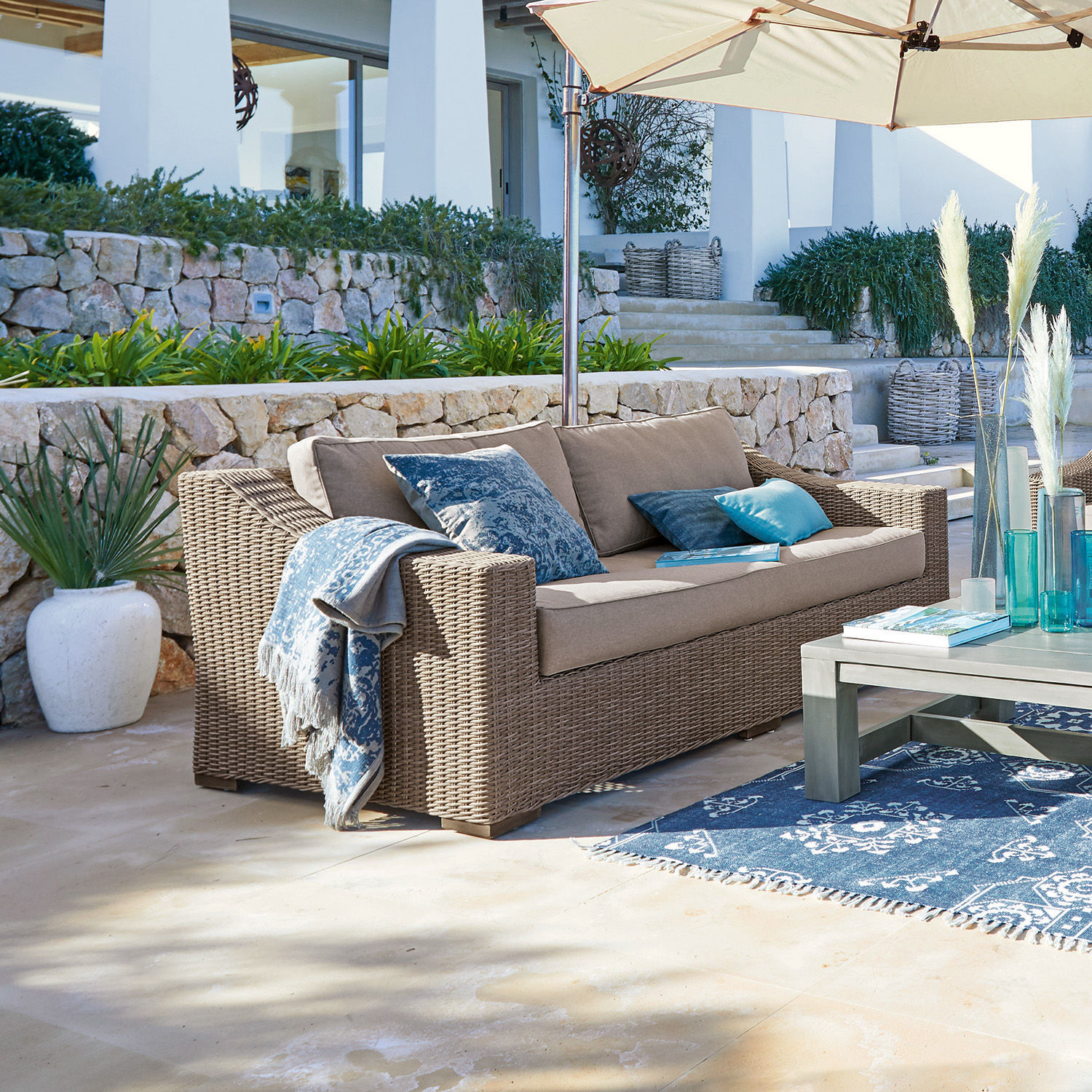 Divano Chestingdale Divano da giardino in rattan beige con cuscini blu e ombrellone su terrazza soleggiata