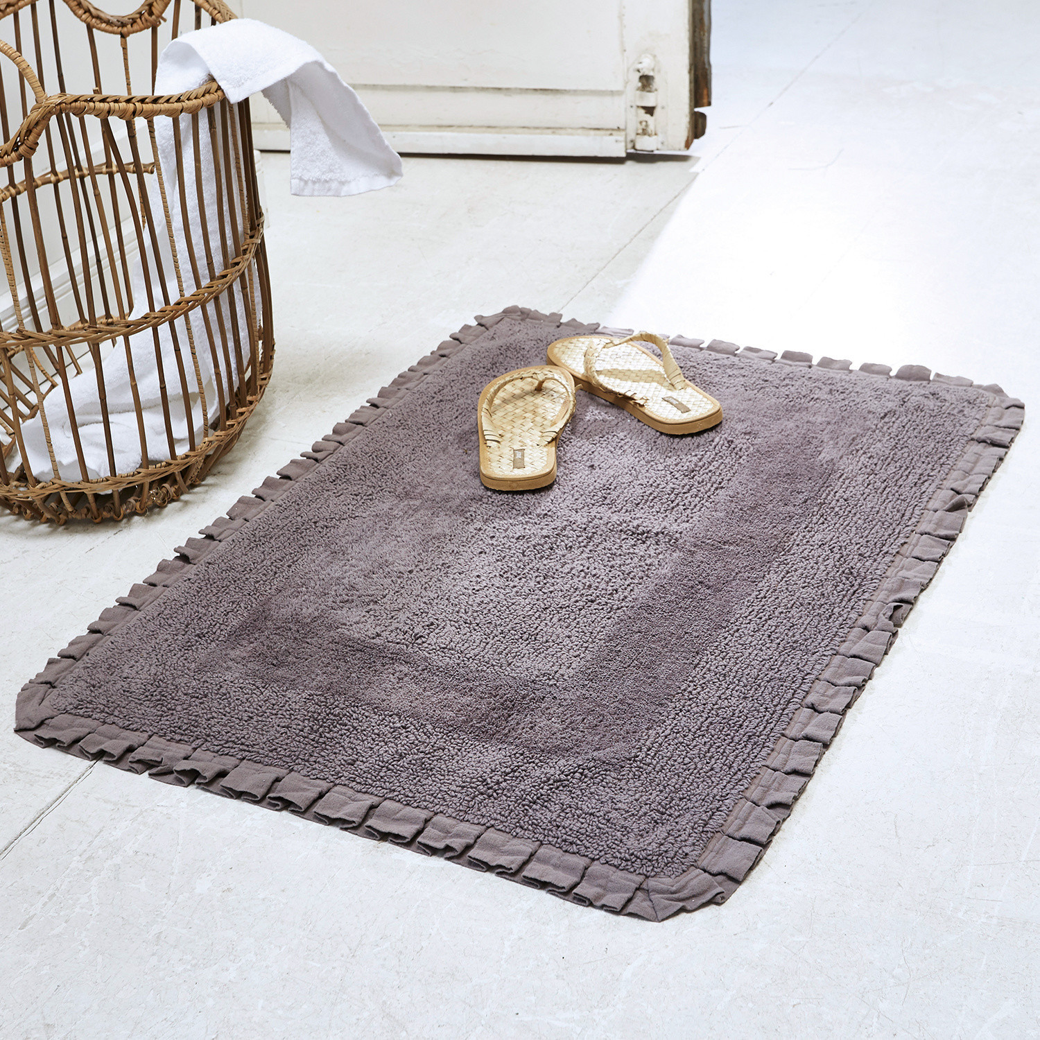 Scendibagno Jasmin Tappeto da bagno grigio in cotone con bordo arricciato su pavimento chiaro con sandali in rattan