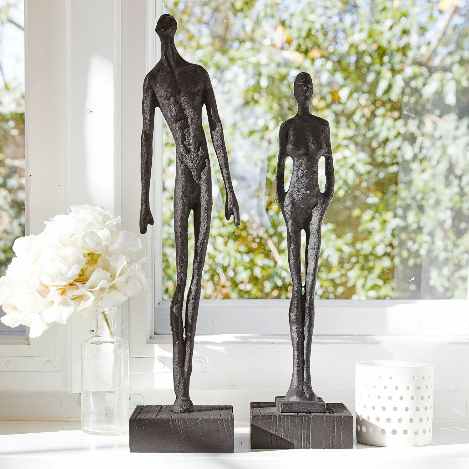 Coppia di statuine Perce Due sculture nere in metallo di figure umane su basi in legno davanti a una finestra luminosa