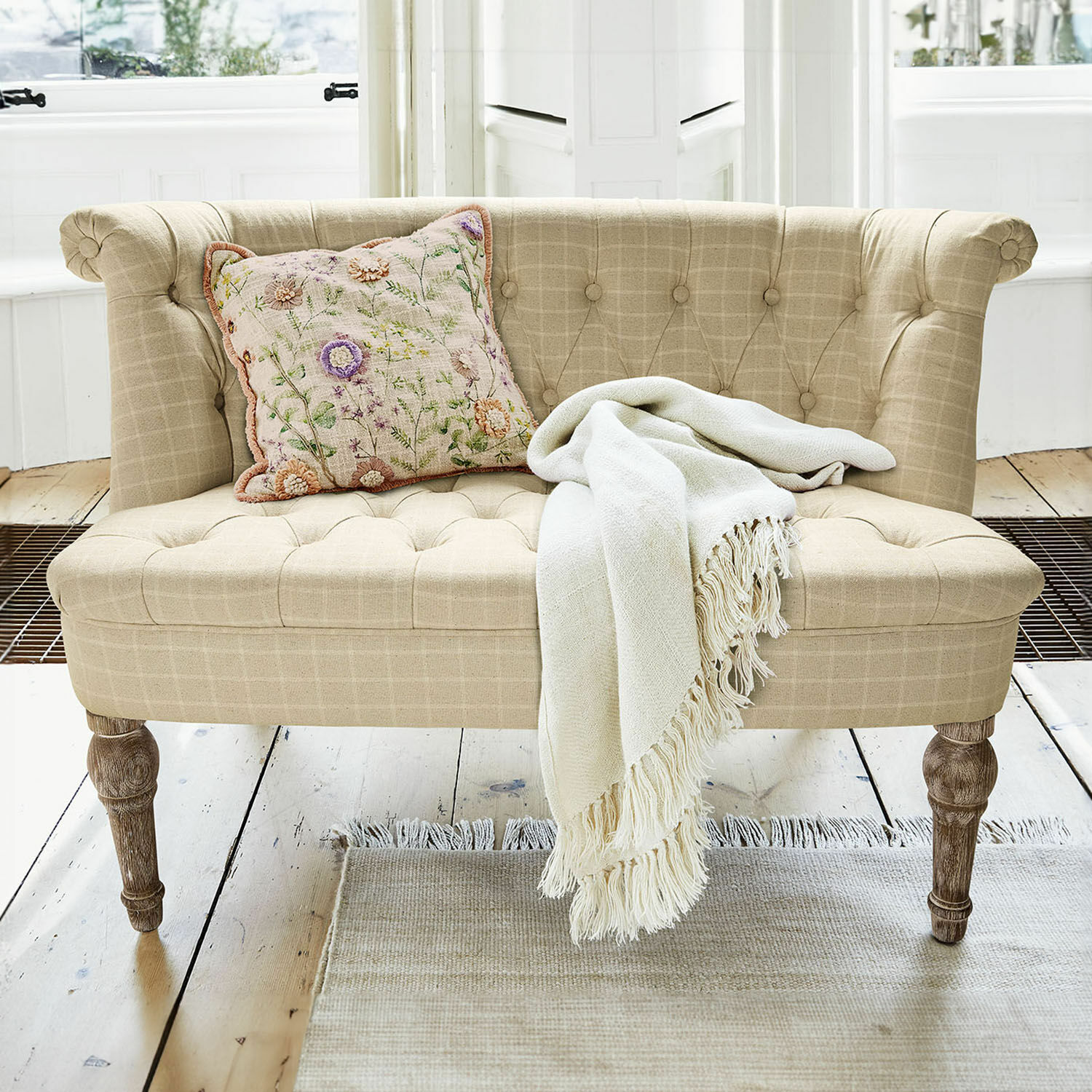 Divano Weralin Divano beige imbottito con gambe in legno, cuscino decorativo e coperta crema su tappeto chiaro
