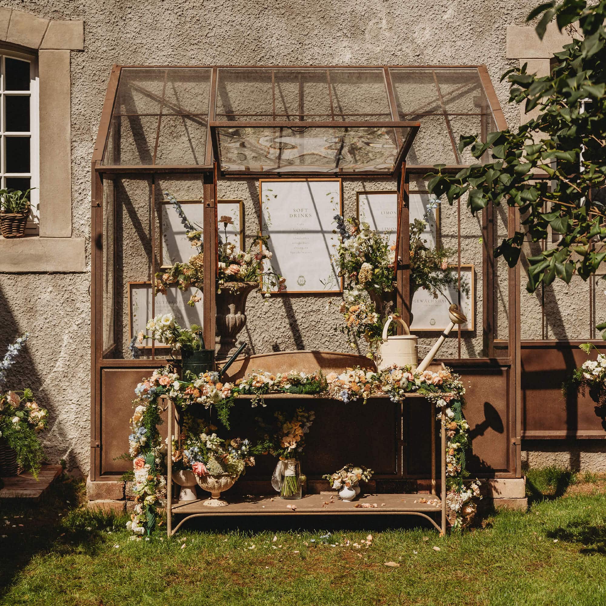 Tavolo da giardino in metallo con decorazioni floreali e annaffiatoi davanti a una serra di vetro
