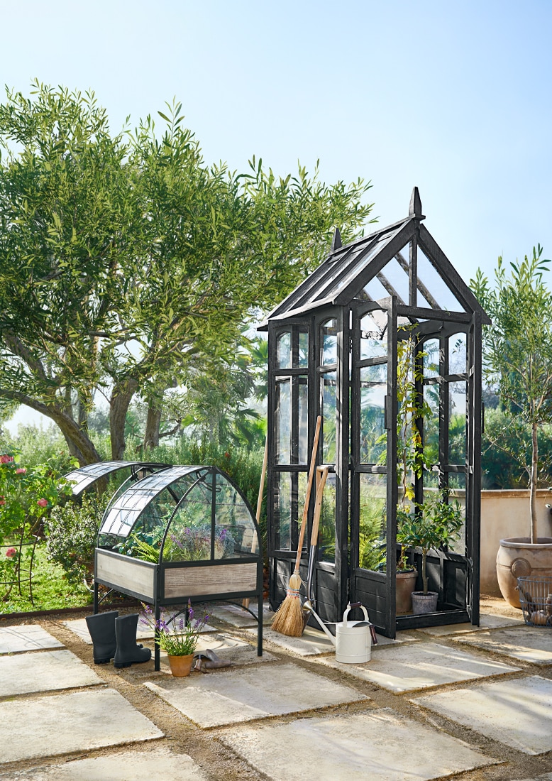 Serra da giardino nera in metallo e vetro con attrezzi da giardinaggio su terrazza soleggiata.