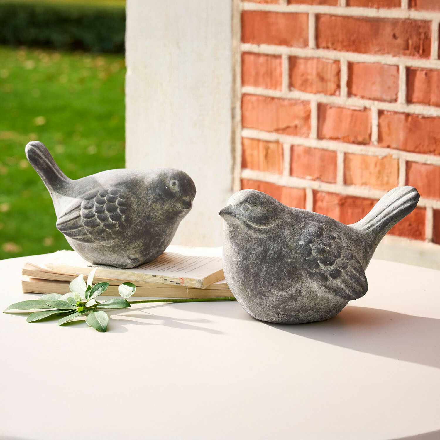 Coppia di uccellini decorativi Haldoríe Due figure decorative di uccelli in pietra grigia su tavolo bianco con libro e foglia verde