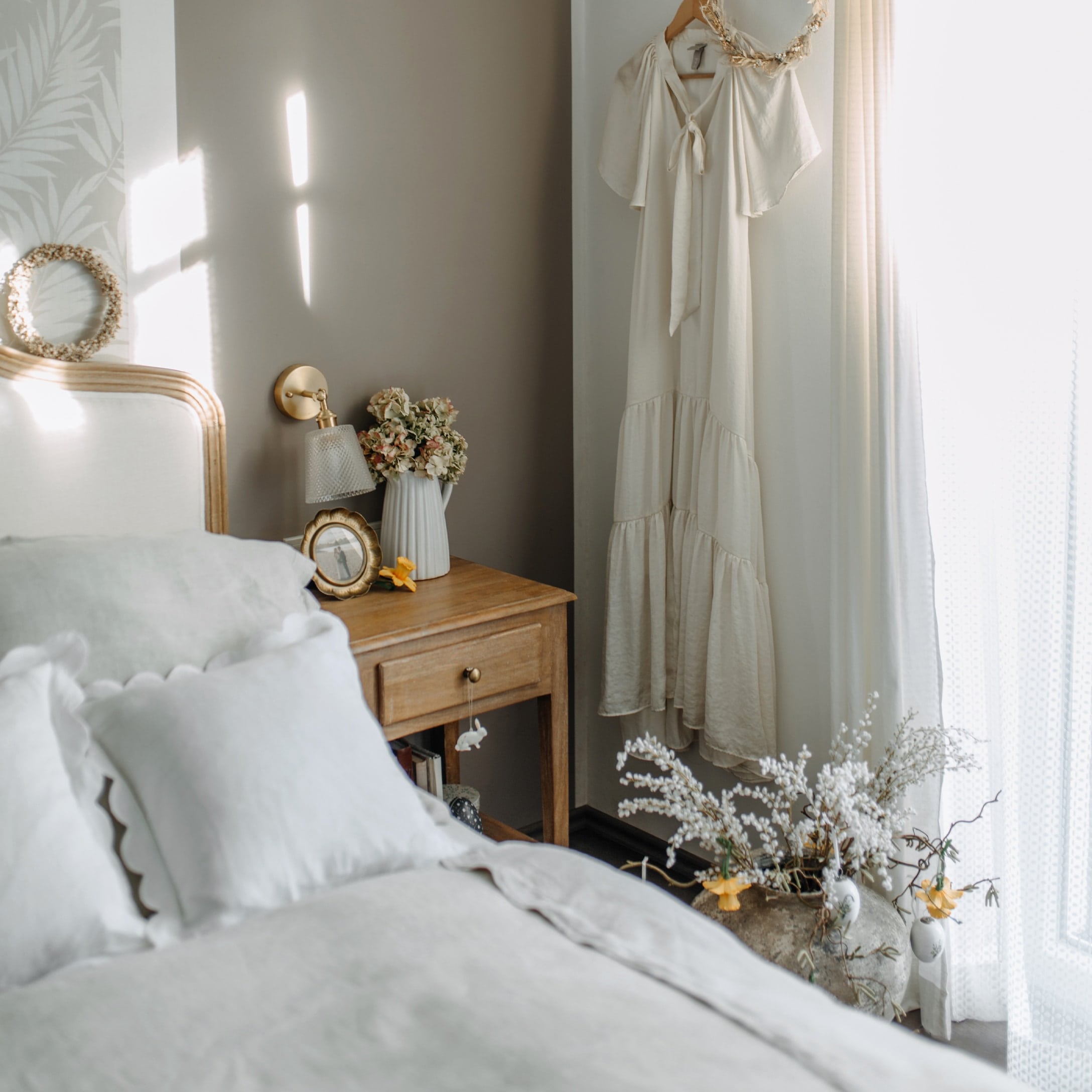 Camera da letto accogliente con comodino in legno, biancheria da letto bianca e vestito crema appeso al muro