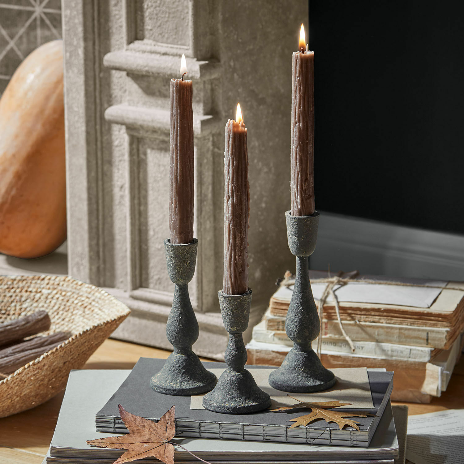 Set di 3 portacandele Lavignoux Tre candelabri in metallo con candele marroni su una pila di libri e una foglia secca
