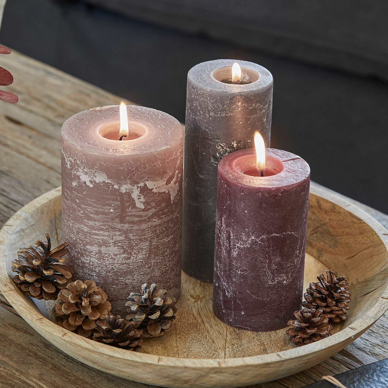 Set di 3 candele Pretin Tre candele accese in tonalità rosa e grigio su vassoio di legno con pigne decorative