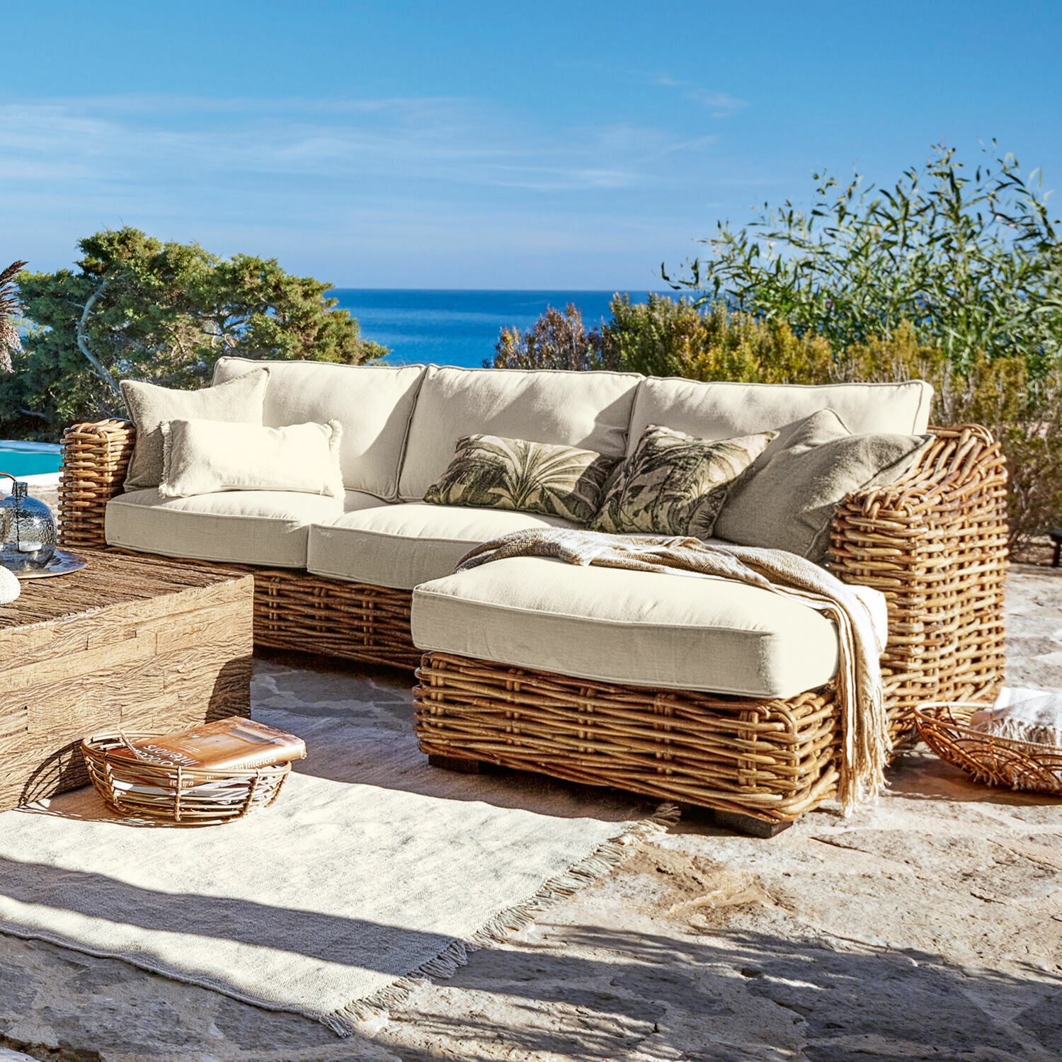 Divano Villedieu Divano angolare in rattan con cuscini crema su terrazza con vista mare, mobili da giardino