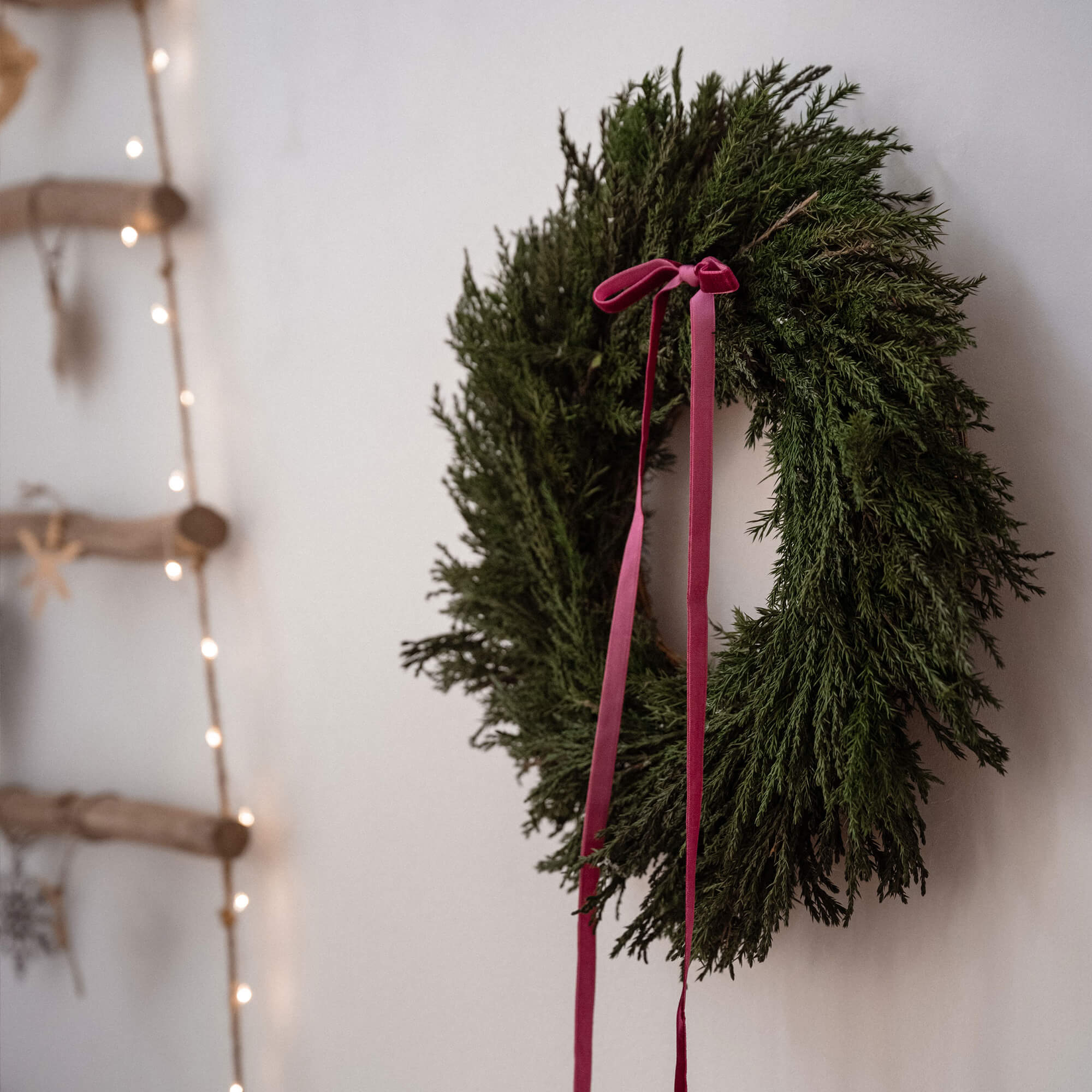 Corona natalizia verde con nastri rossi appesa a parete bianca con luci decorative e legno