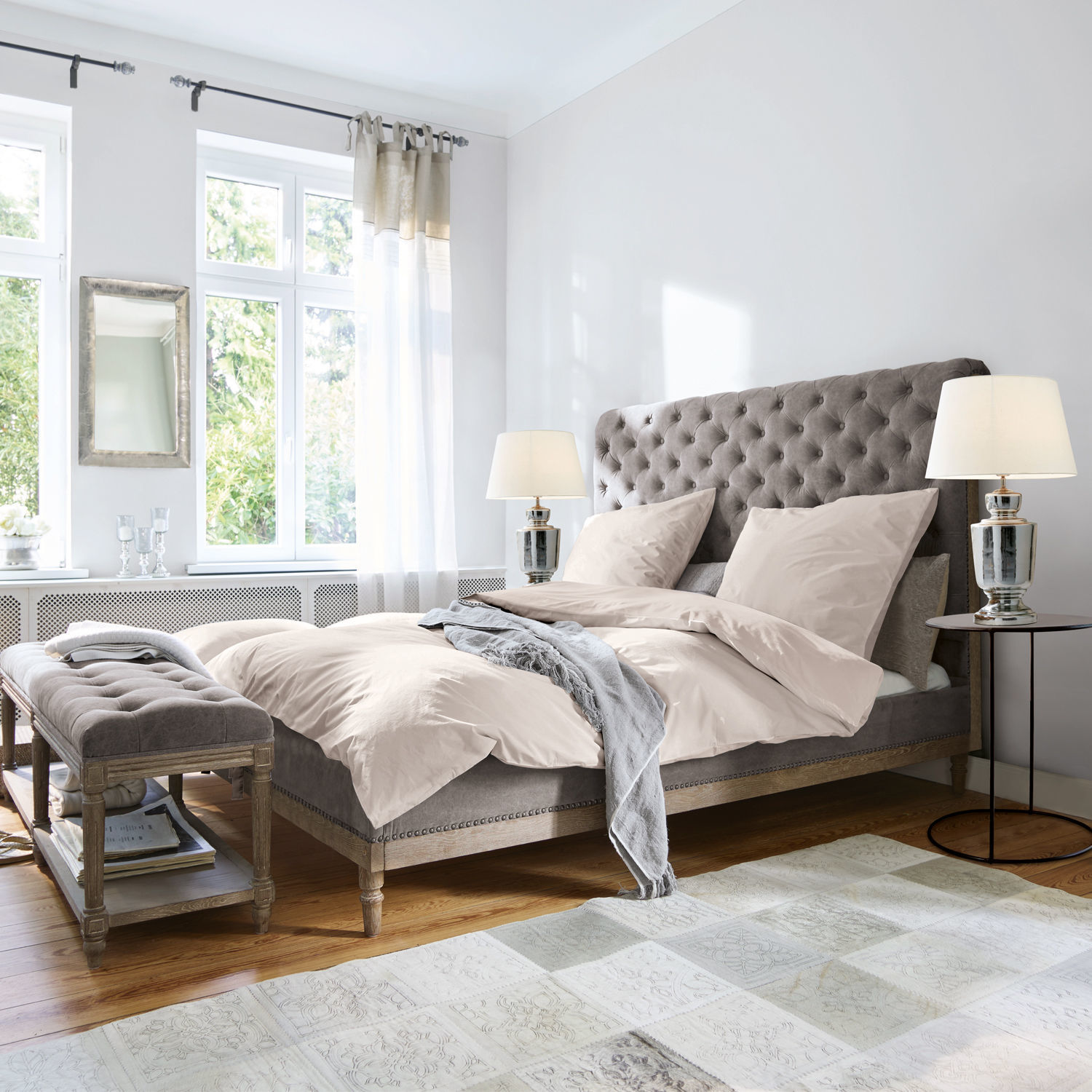 Letto Pozières Letto imbottito grigio con testiera capitonné e biancheria beige in camera luminosa con pavimento in legno