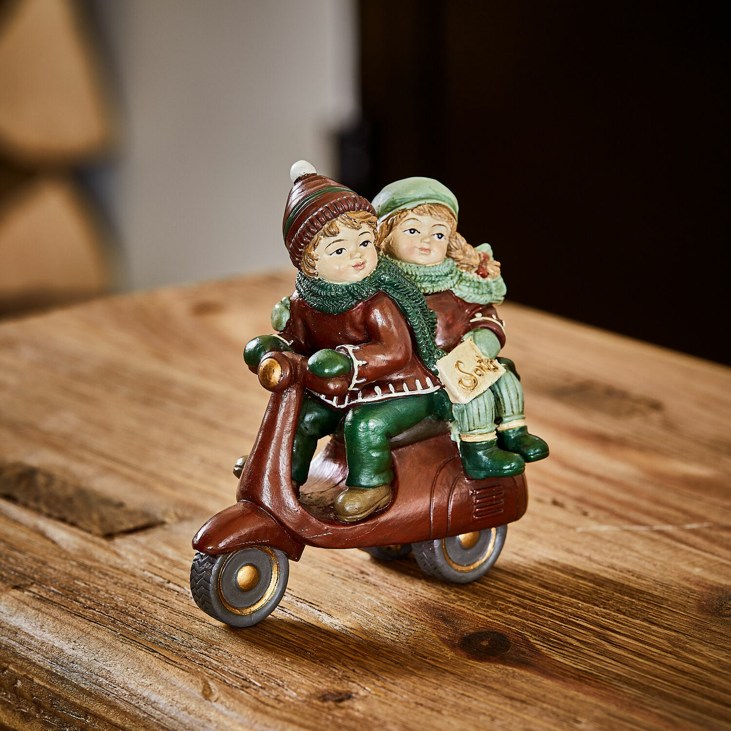Statuina decorativa Dariell Figura di bambini su monopattino marrone con abiti verdi su tavolo in legno decorazione invernale
