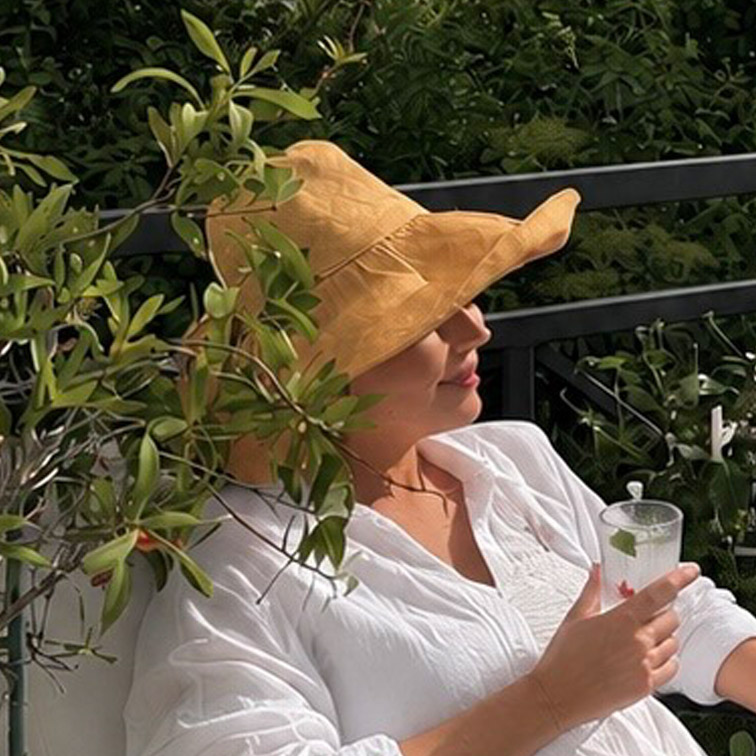 Donna con camicia bianca e cappello di paglia giallo si rilassa in terrazza con bevanda in mano