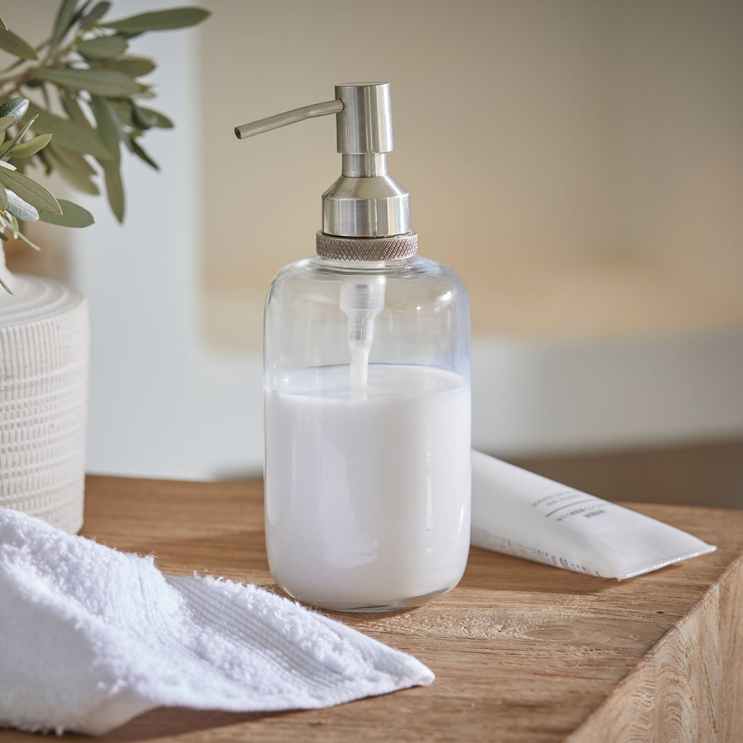 Dispenser di sapone trasparente in acciaio inox con liquido bianco su superficie in legno