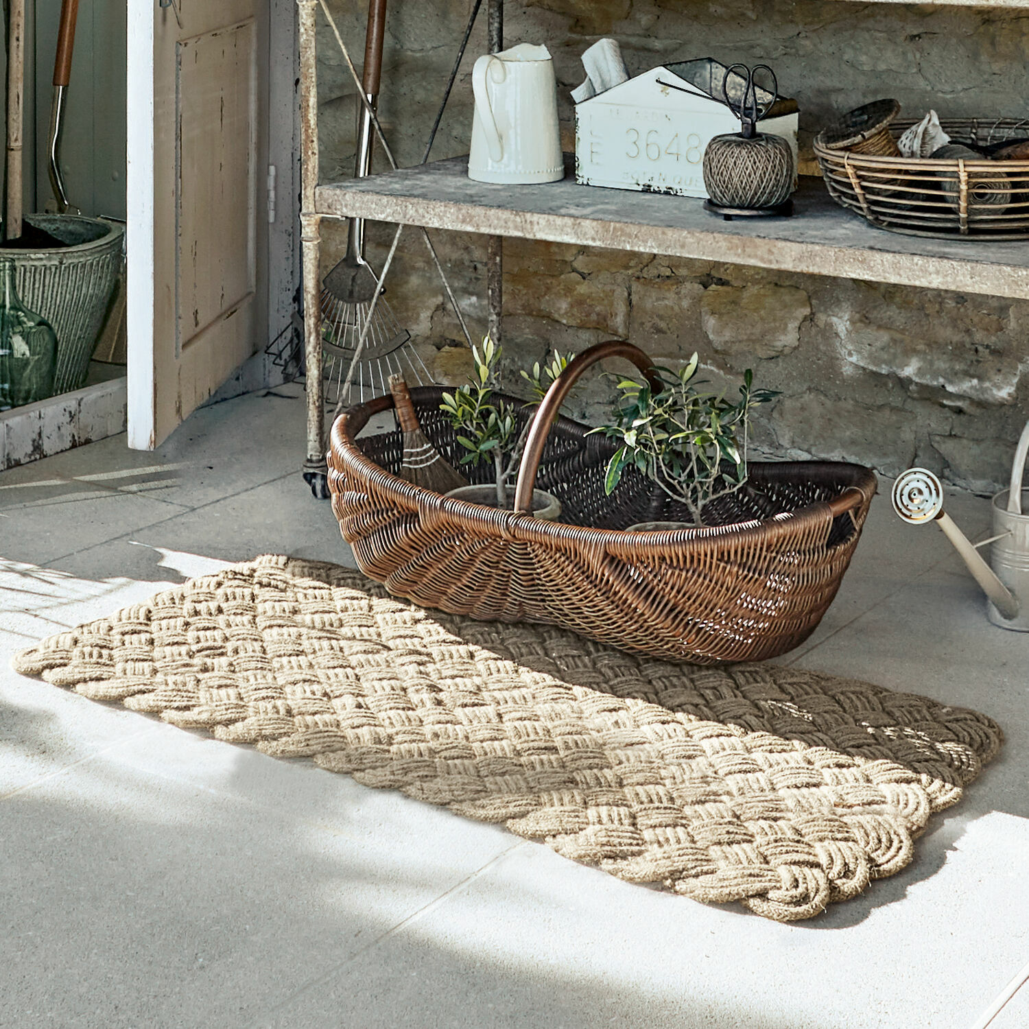 Tappeto Hildas Tappeto beige intrecciato su pavimento in ambiente rustico con cesta in vimini e attrezzi da giardino
