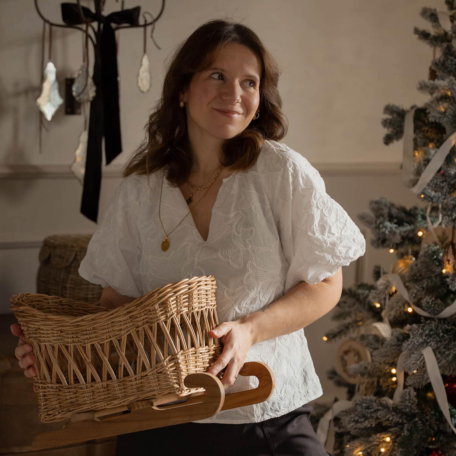 Donna con camicetta bianca tiene una slitta in vimini con base in legno vicino a un albero di Natale decorato