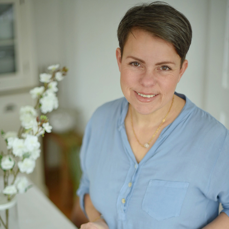 Donna sorridente con camicia azzurra accanto a fiori bianchi in interno luminoso