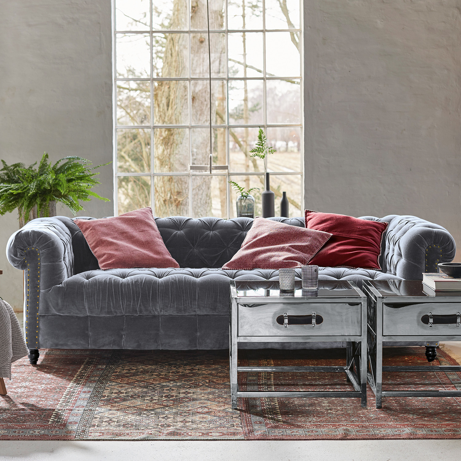 Divano Parpeville Divano Chesterfield in velluto grigio con cuscini rossi e tavolini metallici su tappeto decorativo