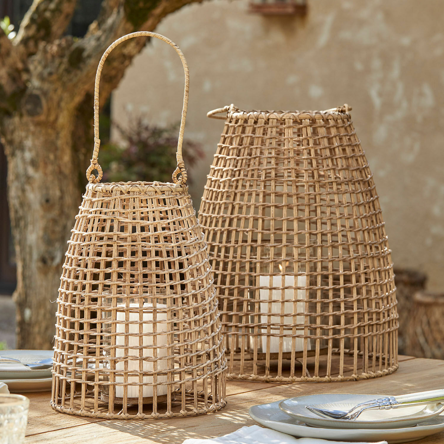 Coppia di lanterne Hillendale Due lanterne in rattan naturale con candele bianche su un tavolo di legno all'aperto