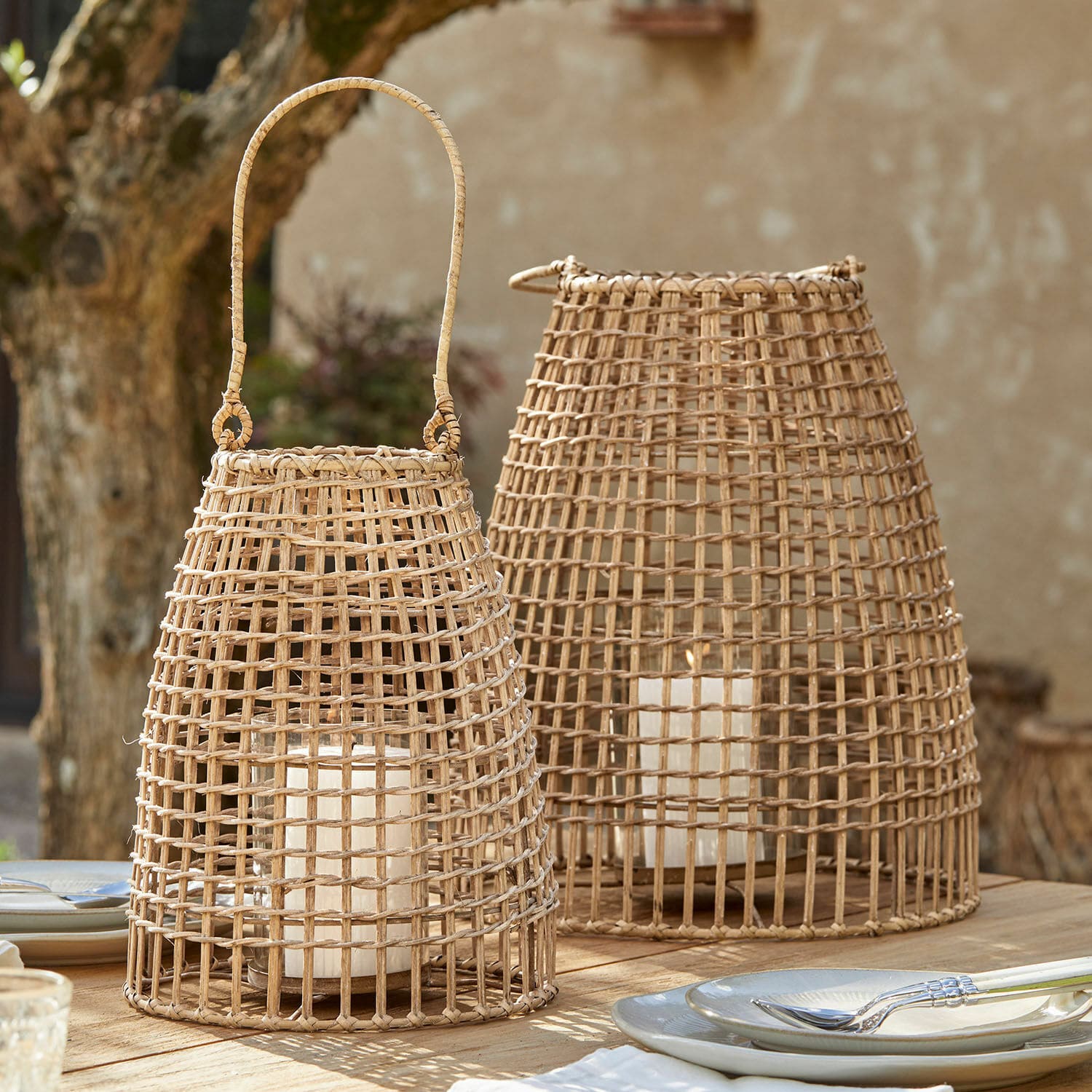 Coppia di lanterne Hillendale Due lanterne in rattan naturale con candele bianche su un tavolo di legno all'aperto