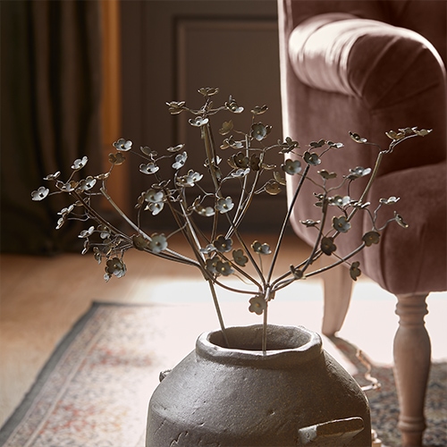Ramo decorativo in metallo con fiori in vaso di ceramica grigia su tappeto accanto a poltrona rosa