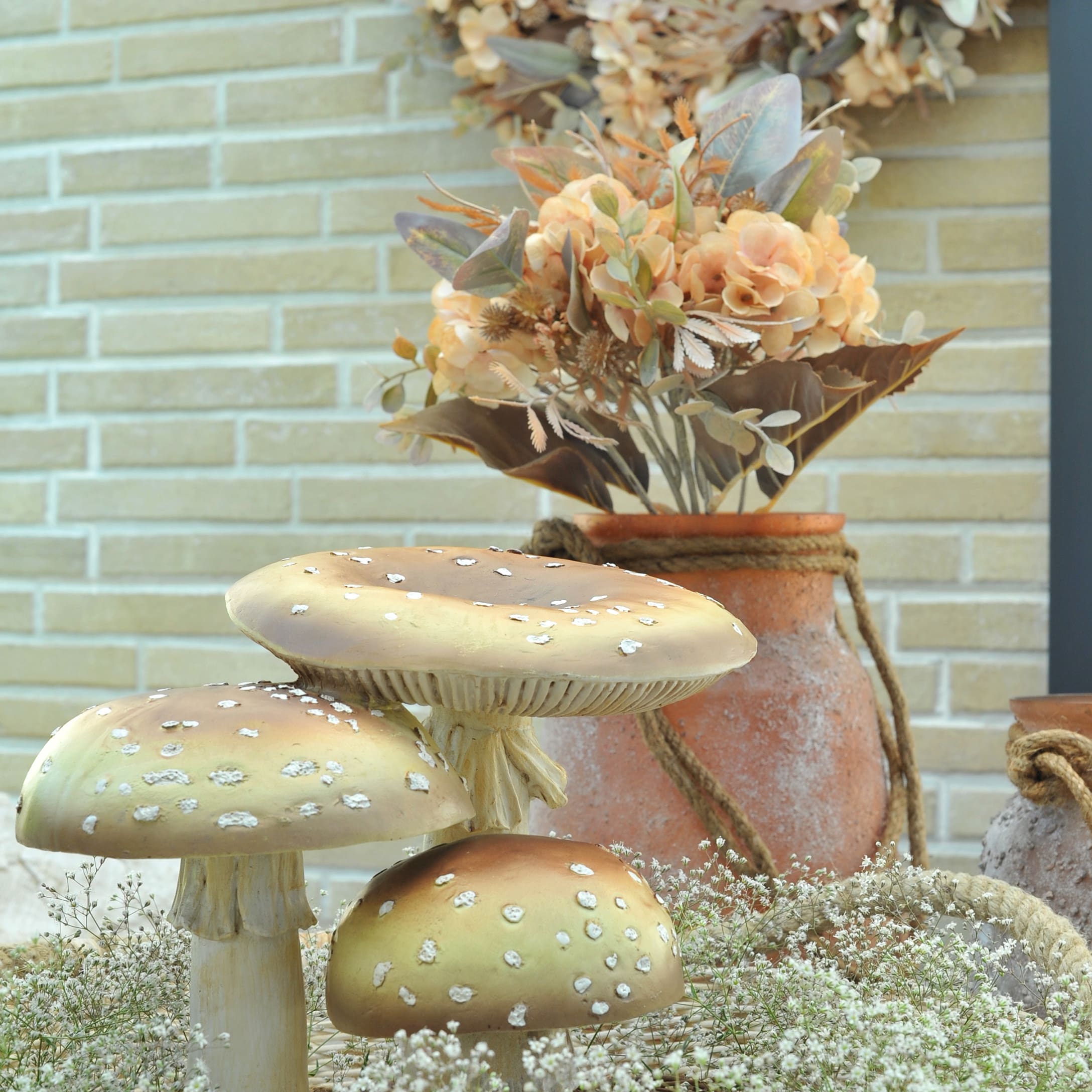 Funghi decorativi beige e marroni con vaso in terracotta e fiori autunnali davanti a muro di mattoni