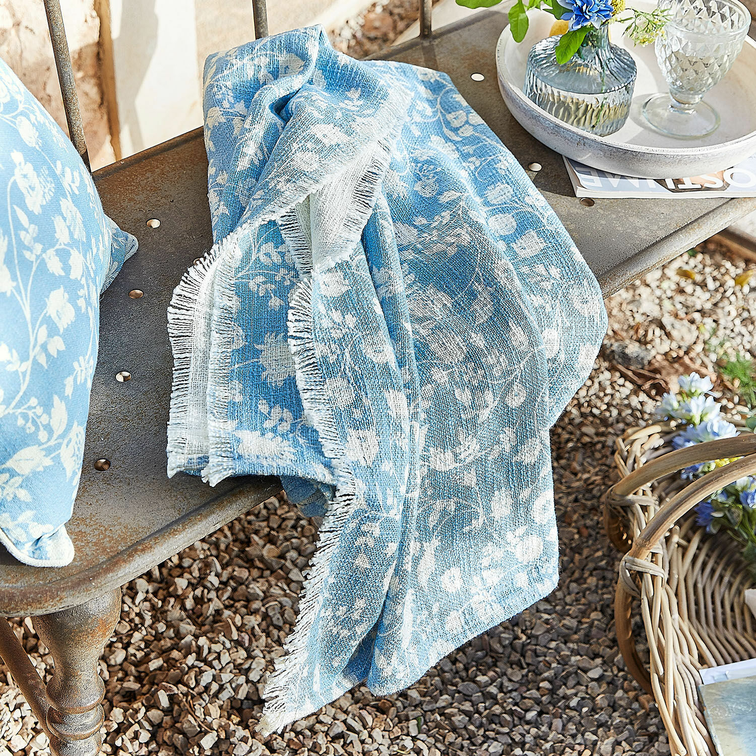Plaid Corvelique Coperta di cotone azzurra con motivo floreale bianco su panca in metallo in giardino