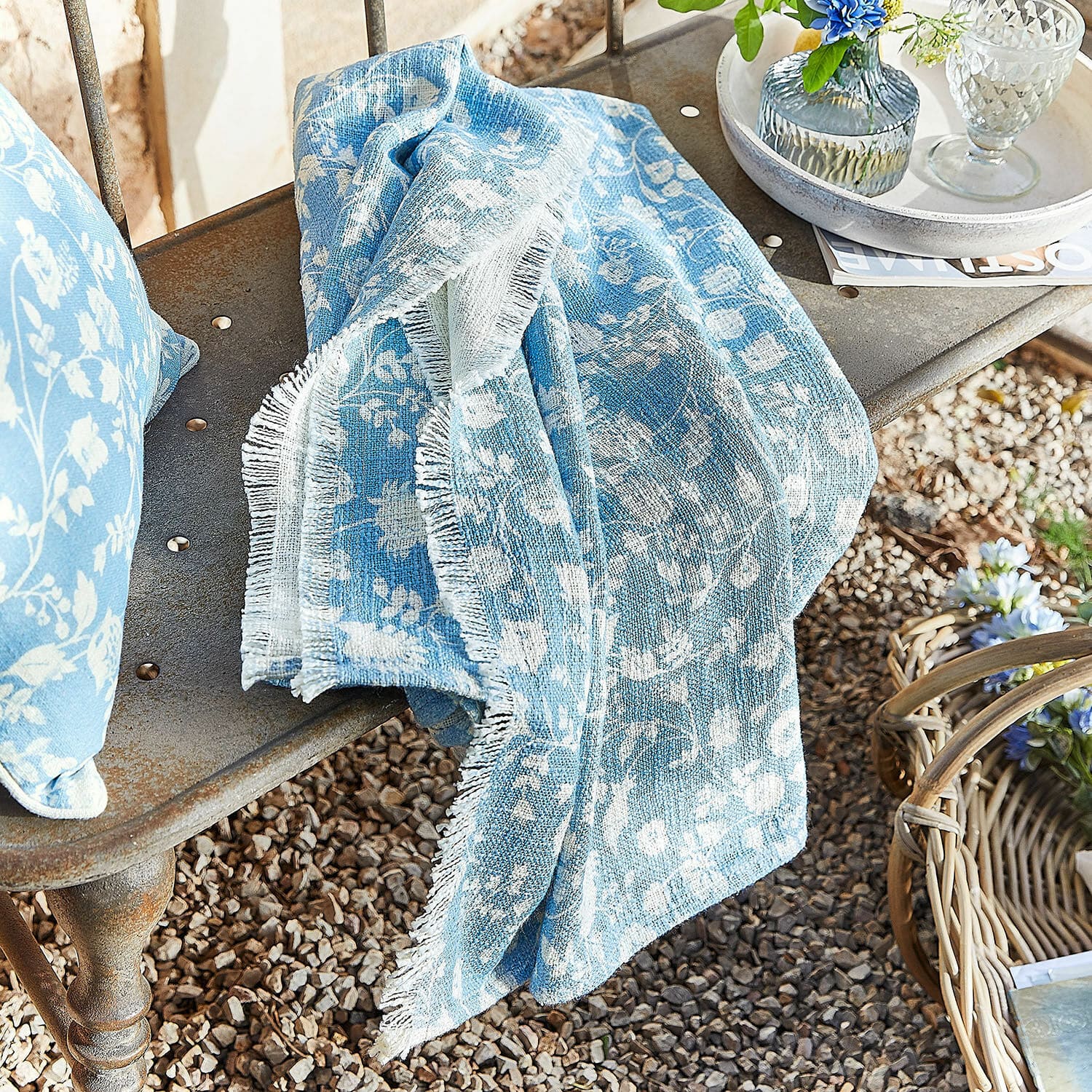 Plaid Corvelique Coperta di cotone azzurra con motivo floreale bianco su panca in metallo in giardino