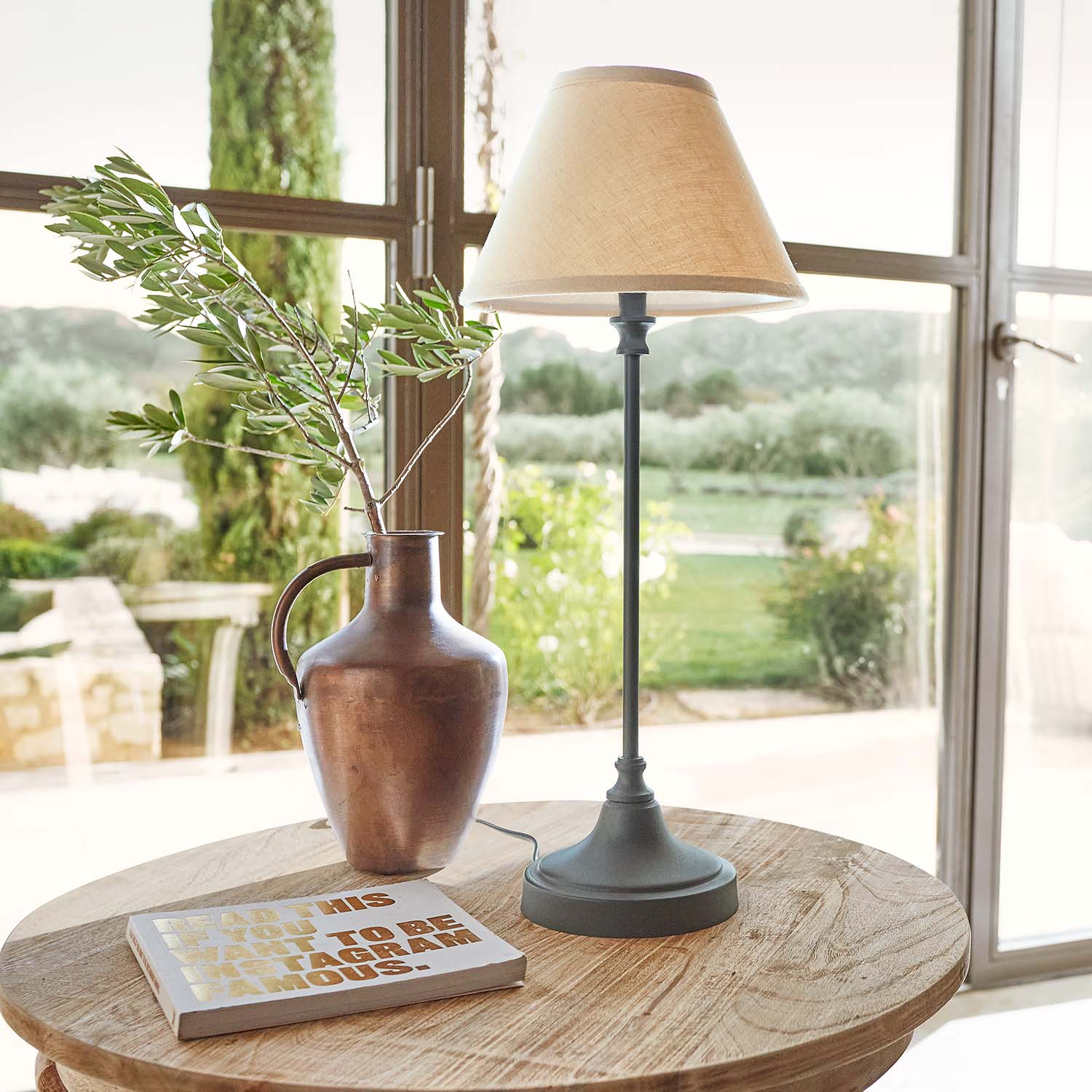 Brocca in metallo color rame con ramo e lampada da tavolo con paralume beige su tavolo in legno