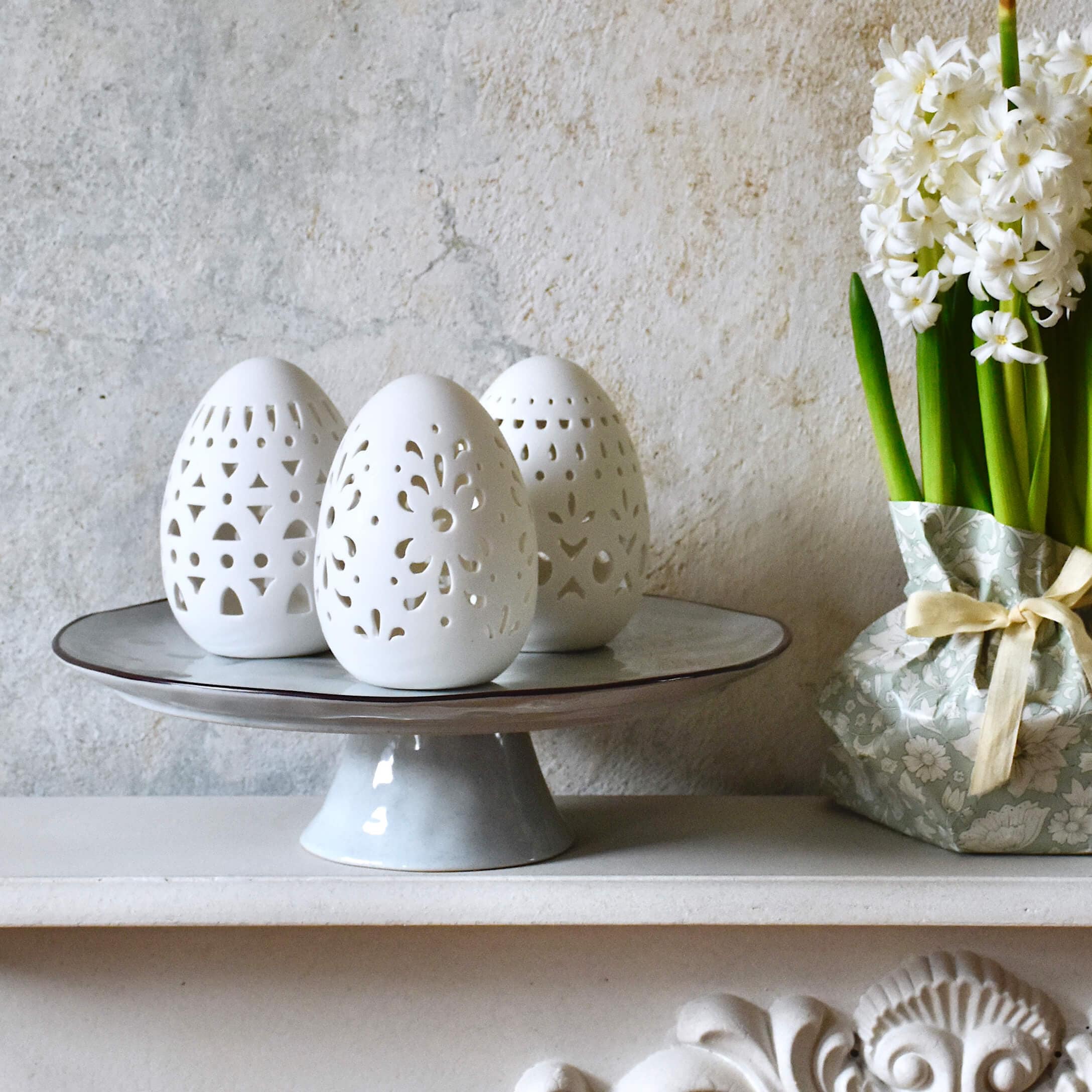 Uova di ceramica bianca traforata su piatto grigio accanto a fiori bianchi in confezione decorativa