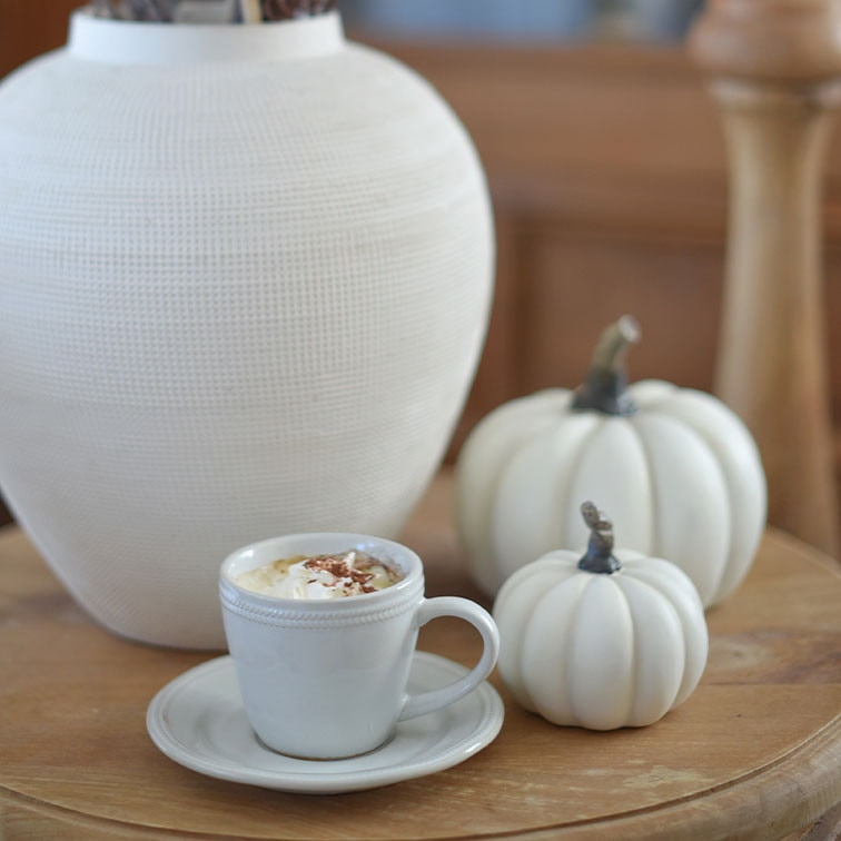 Tazza in ceramica bianca con caffè e panna su tavolo in legno accanto a zucche decorative bianche e vaso