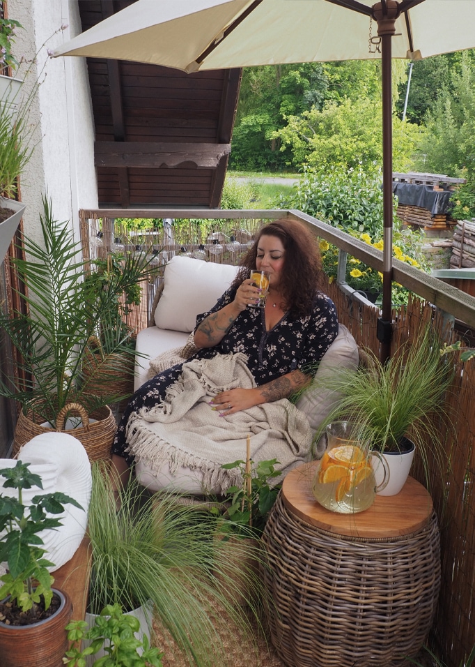 Donna che si rilassa su balcone con tavolo in vimini e coperta beige, circondata da piante verdi.