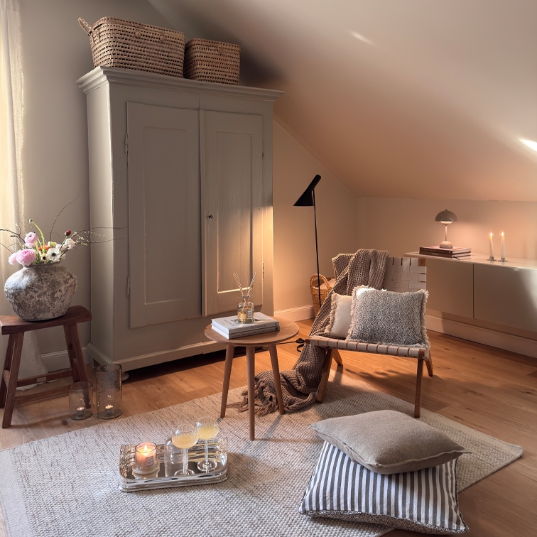 Soggiorno accogliente con poltrona beige, tavolino in legno e armadio sotto il tetto inclinato, decorazioni e lampade