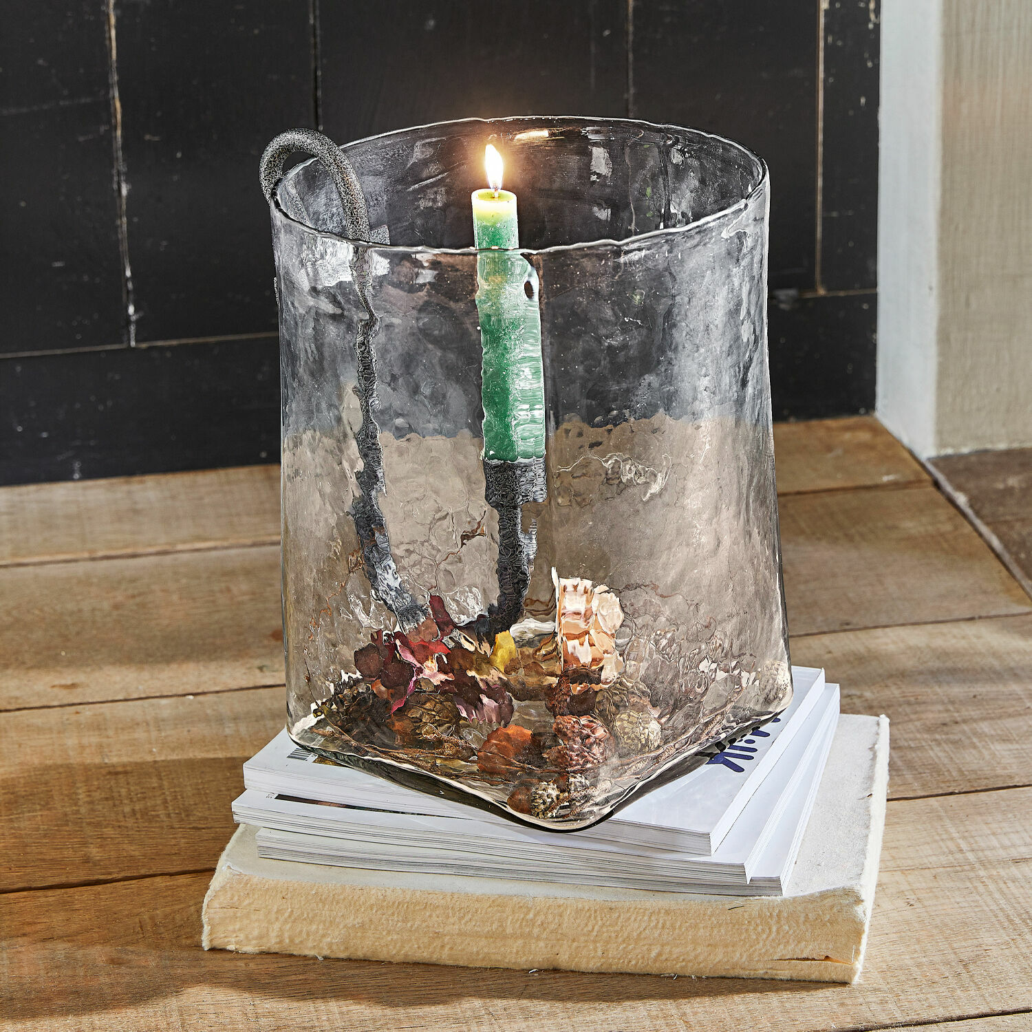 Portacandele in vetro con manico in metallo e candela verde su tavolo in legno con decorazioni di pigne