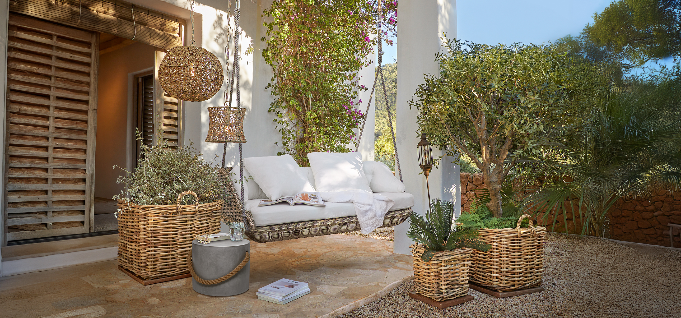 Dondolo in rattan con cuscini bianchi su terrazza con cesti di vimini e piante verdi