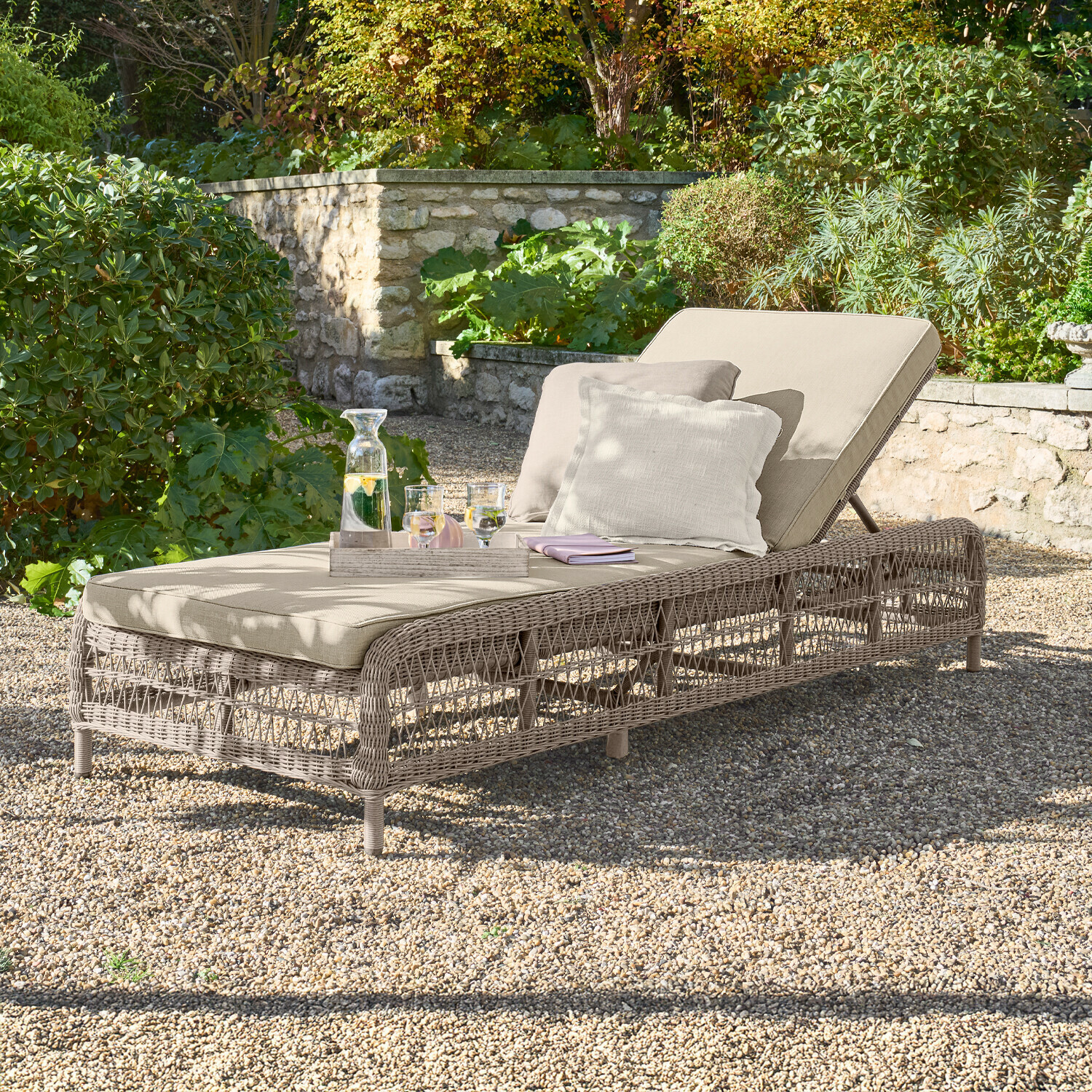 Lettino Hereford Lettino da giardino beige con cuscino e struttura intrecciata su ghiaia in giardino verde