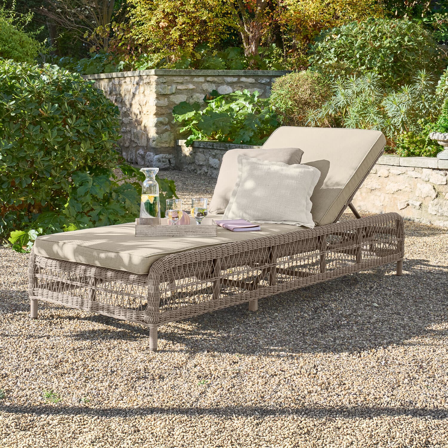 Lettino Hereford Lettino da giardino beige con cuscino e struttura intrecciata su ghiaia in giardino verde