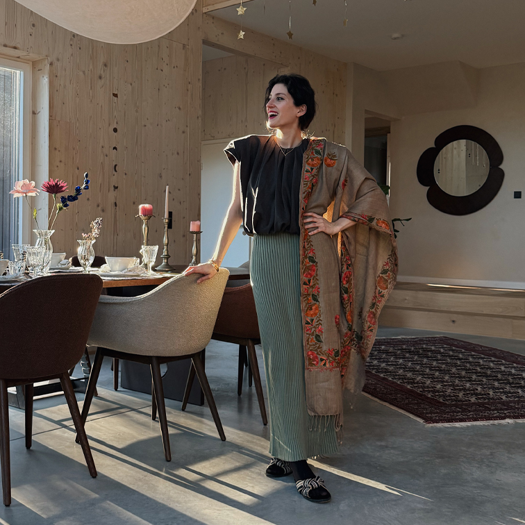 Donna con gonna plissettata verde, top nero e sciarpa beige con motivo floreale in interno moderno
