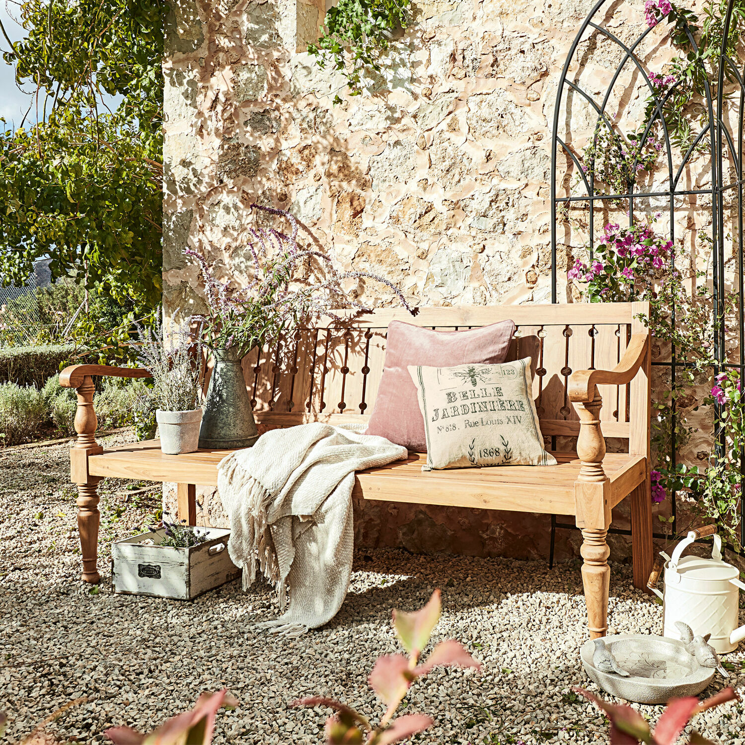 Panca Loudoun Panca da giardino in legno chiaro con cuscini e coperta su patio di ghiaia con piante e decorazioni