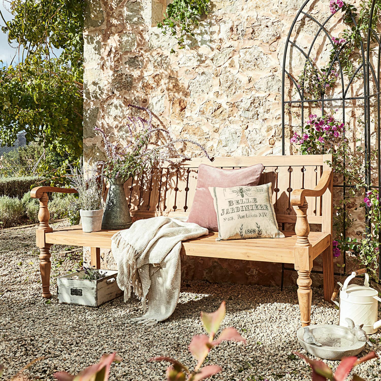 Panca Loudoun Panca da giardino in legno chiaro con cuscini e coperta su patio di ghiaia con piante e decorazioni
